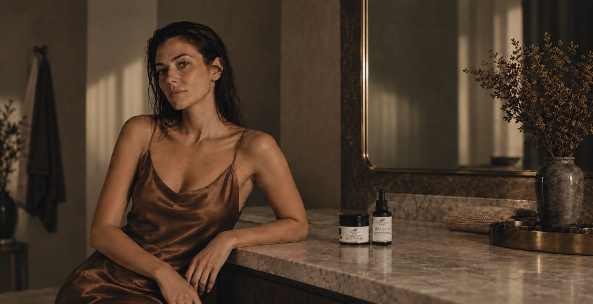 A woman with dark hair sits at a marble bathroom counter wearing a bronze satin slip dress