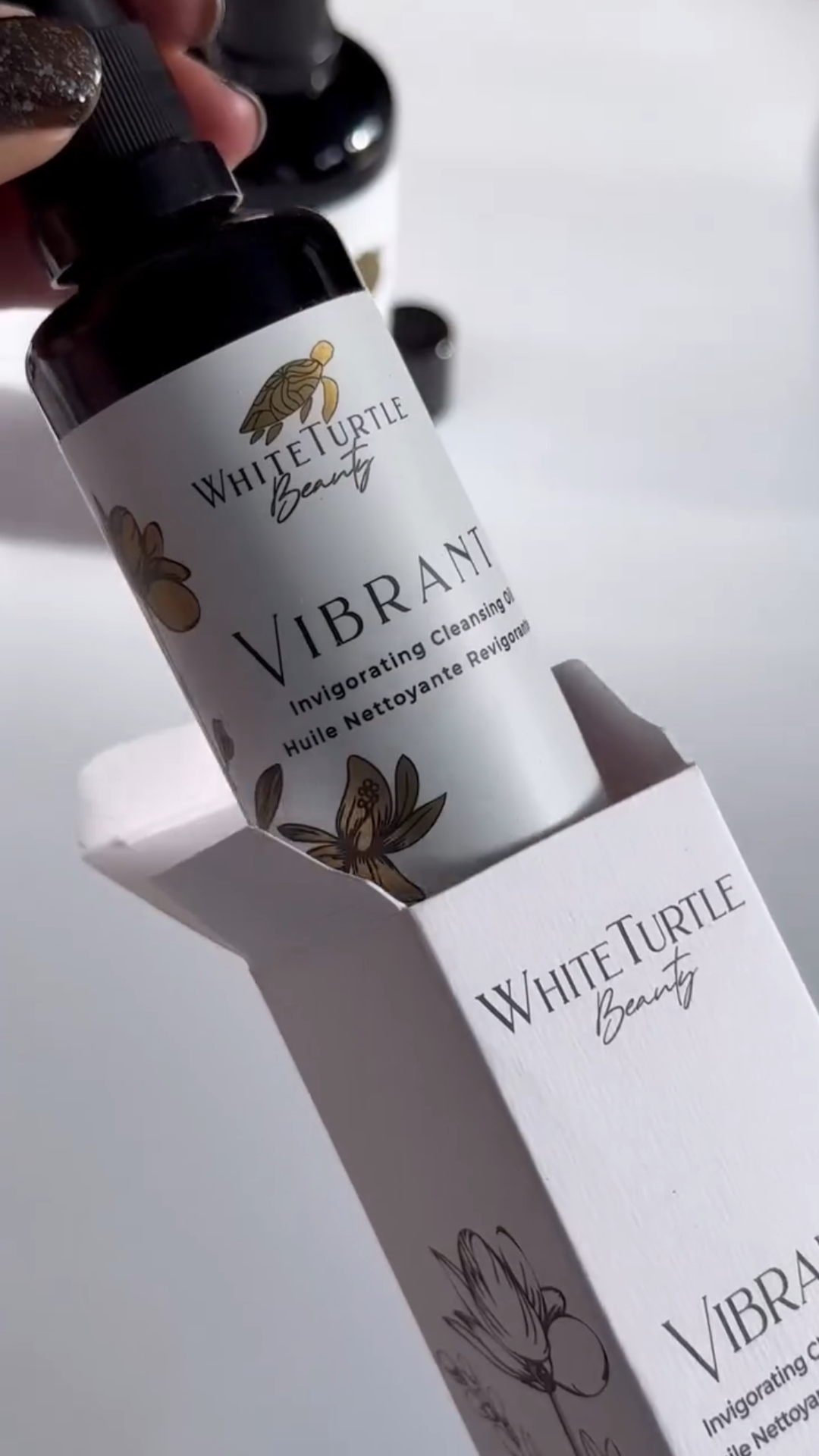 A hand holds a dark bottle labeled 'White Turtle Beauty Vibrant Invigorating Cleansing Oil' 