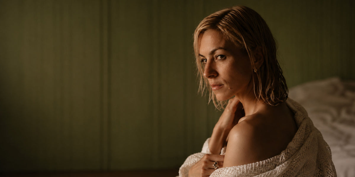 A woman with wet, shoulder-length blonde hair sits wrapped in a white knit blanket 