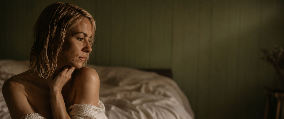 A woman wrapped in a white towel sits on a bed in a dimly lit room with green walls.