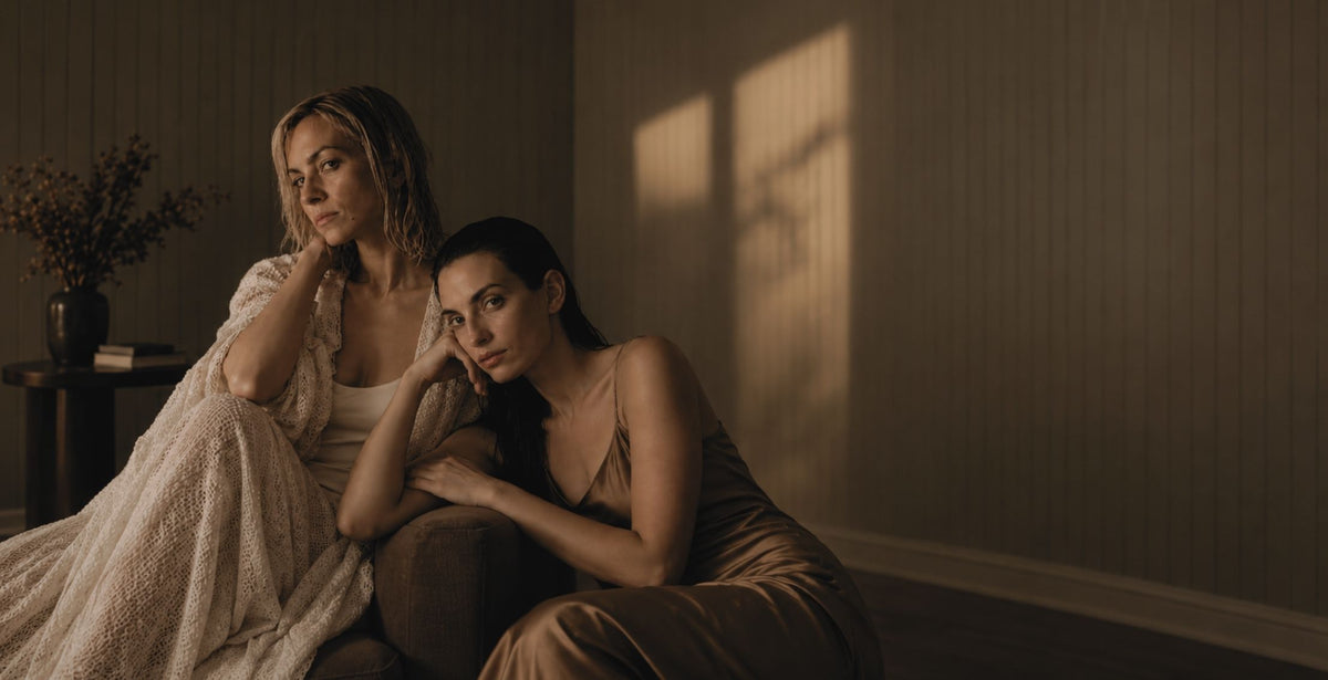 Two women sitting together in a dimly lit room with warm sunlight casting shadows on the wall